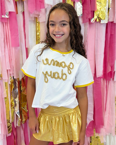 Young girl wearing a white shirt with gold text and a gold skirt in front of pink and gold draped fabric.