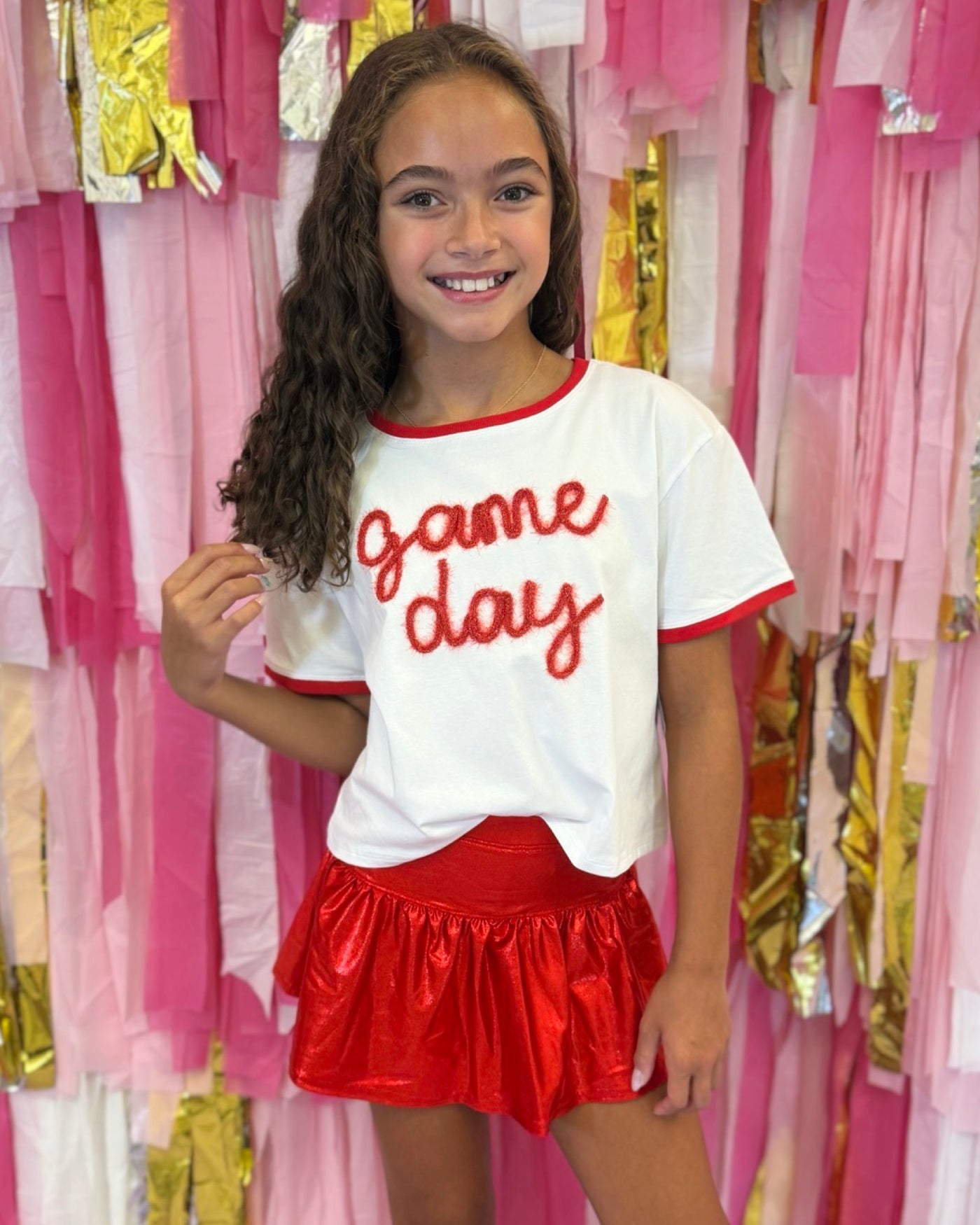 Tween girl wearing a 'Game Day' shirt and red skirt in front of pink and gold streamers.