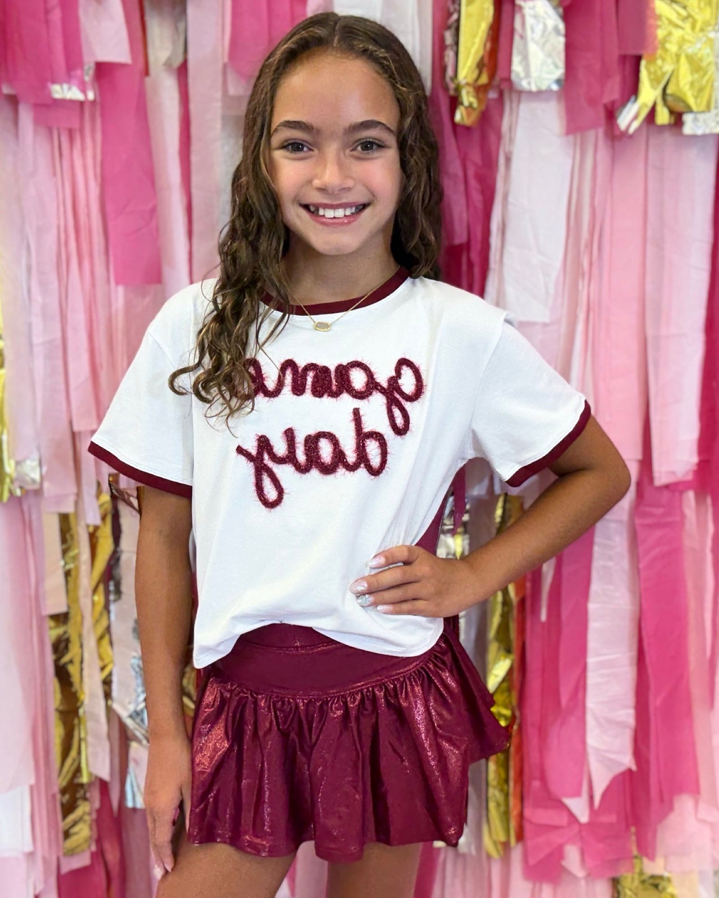 Tween girl wearing a white t-shirt with red text and maroon skirt in front of pink and gold curtains.